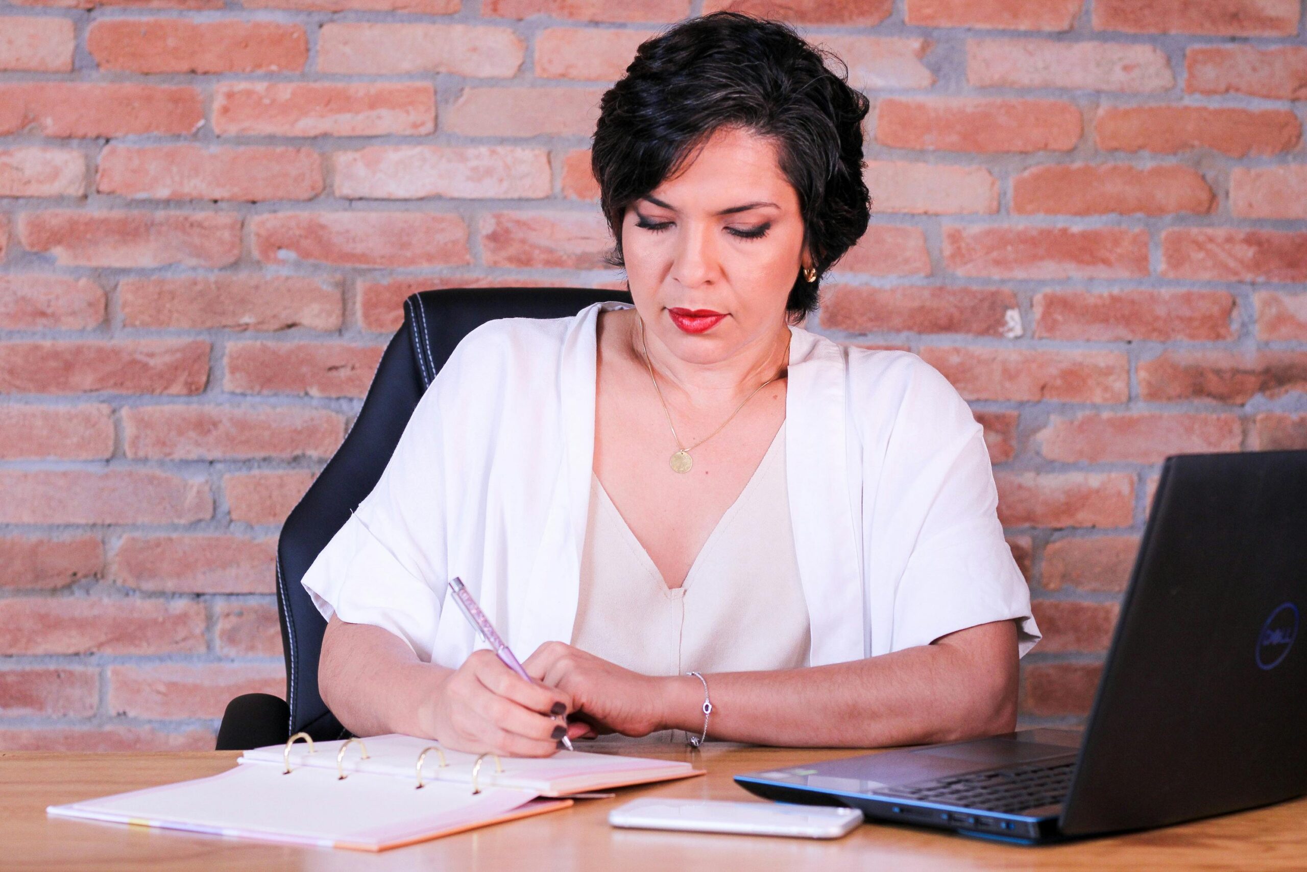 Elegant businesswoman writing in notebook at modern office desk with laptop.